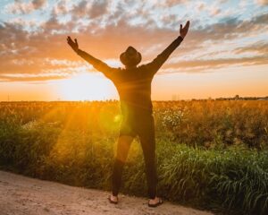 Frei atmender Mann mit erhobenen Armen im Sonnenuntergang, stehend vor einer grünen Wiese