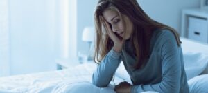 Erschöpfte Frau sitzt morgens im Bett und hält sich den Kopf – Symbolbild für Fatigue und chronische Müdigkeit