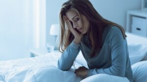 Erschöpfte Frau sitzt morgens im Bett und hält sich den Kopf – Symbolbild für Fatigue und chronische Müdigkeit